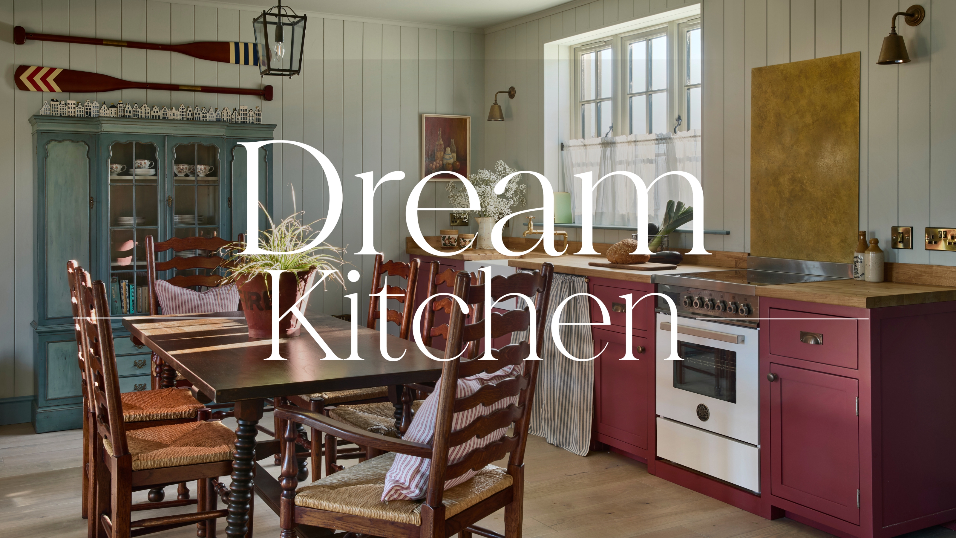 Eine schottische Küsten-Cottage-Küche mit sanft blau getäfelten Wänden, burgunderfarbenen Schränken, einem hölzernen Küchentisch und einem dunkelblauen antiken Schrank