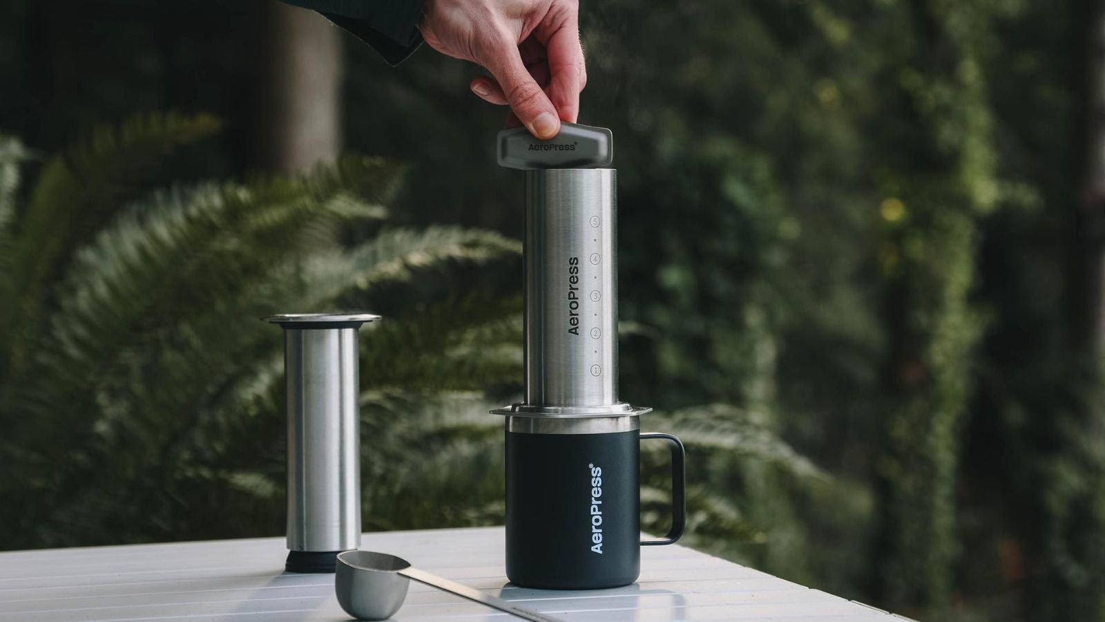 Ein AeroPress-Werkzeug aus Edelstahl auf einem schwarzen Reisebecher, der von den Fingern einer anderen Person auf einem weißen Tisch festgehalten wird. Grüne Blattpflanzen im Hintergrund unscharf. 