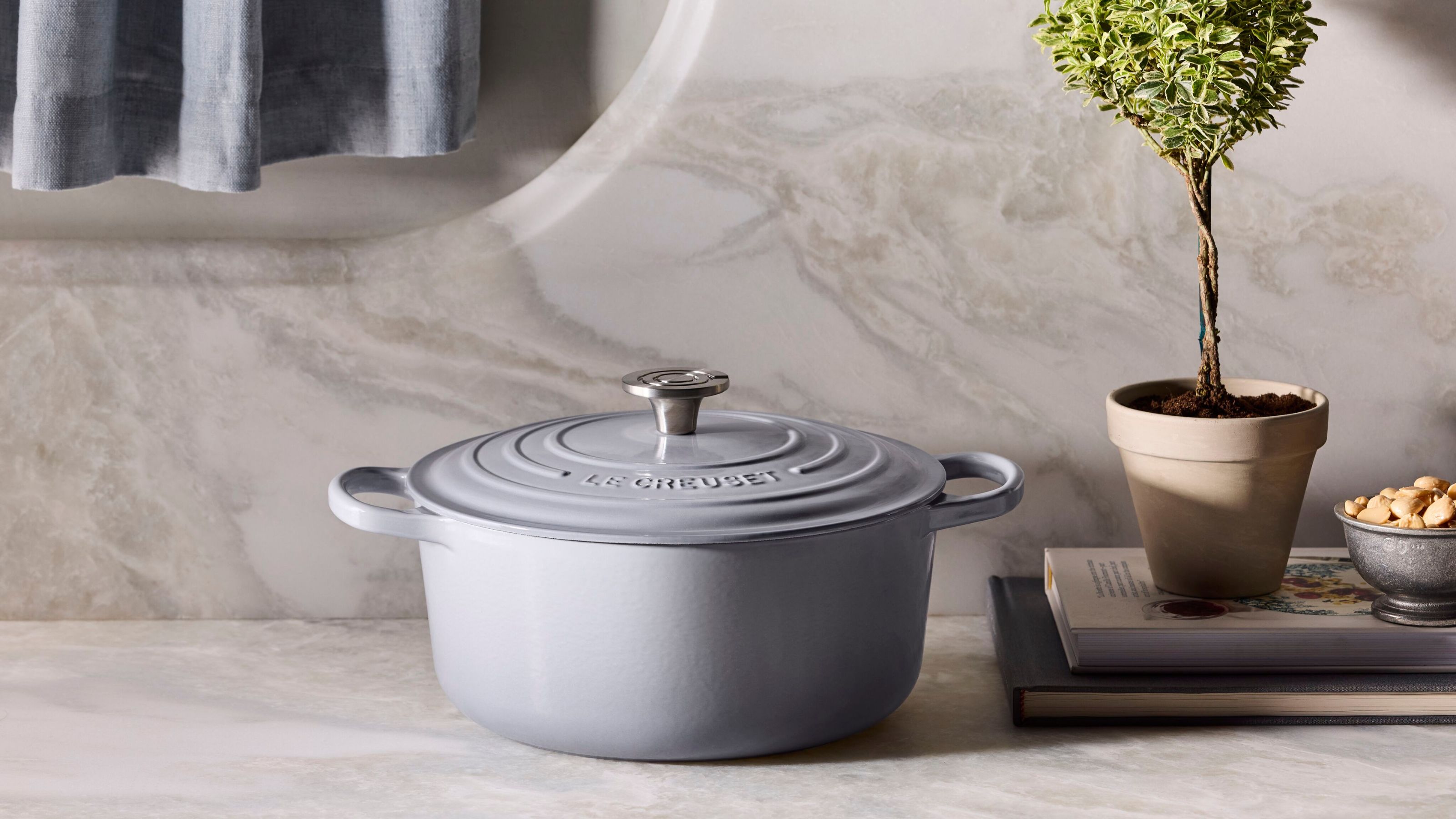 Ein holländischer Ofen von Le Creuset in sanftem Taubengrau vor einem marmorierten Hintergrund neben einem kleinen Baum im Topf 