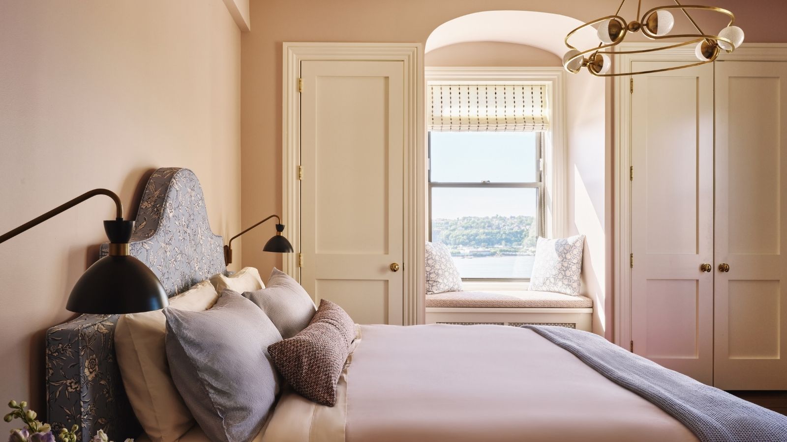 Ein beruhigendes Schlafzimmer in zartem Rosa mit cremefarbenen Einbauschränken und einem Bogenfenster mit Sitzecke. Ein großes Bett mit lila Bettwäsche, blauen Kissen und zwei schwarzen Wandlampen.