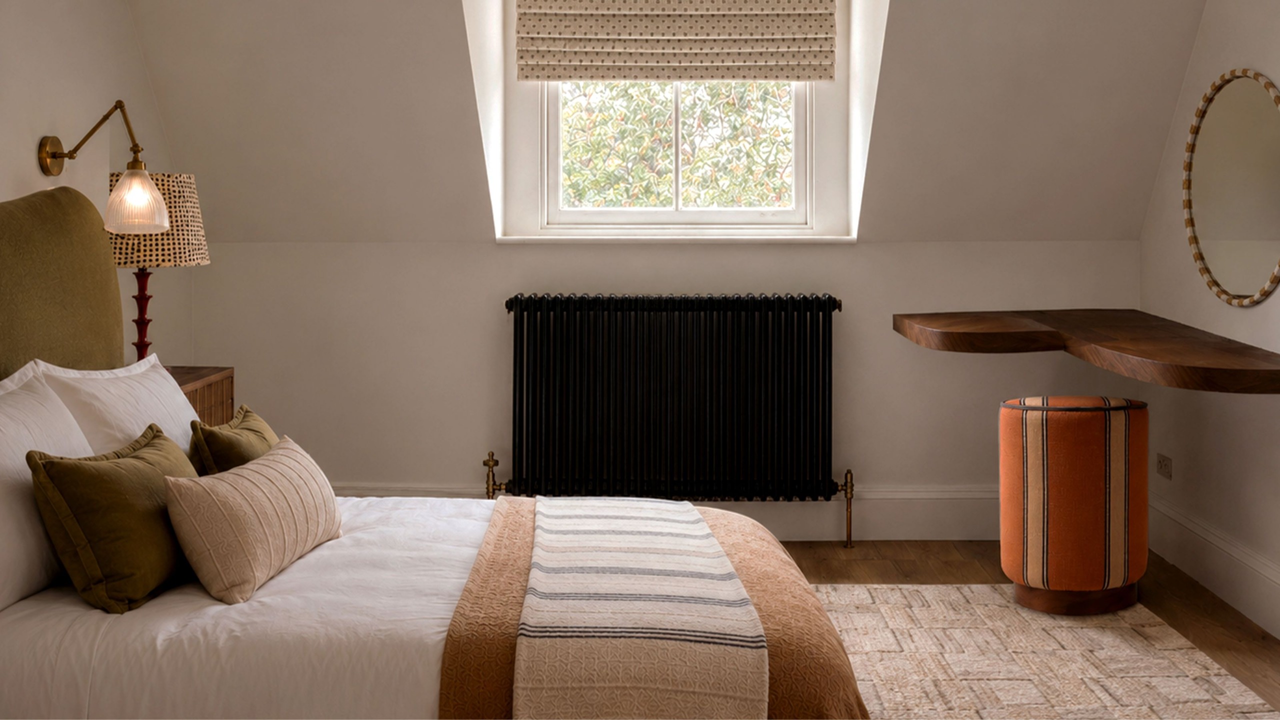 Schlafzimmer mit weißen Wänden, einem Fenster mit einem Raffrollo in der Mitte, einem olivgrünen Bett mit neutraler Bettwäsche und einer Wandleuchte darüber, einem cremefarbenen Teppich und einem orangefarbenen Sitzkissen in der Ecke.