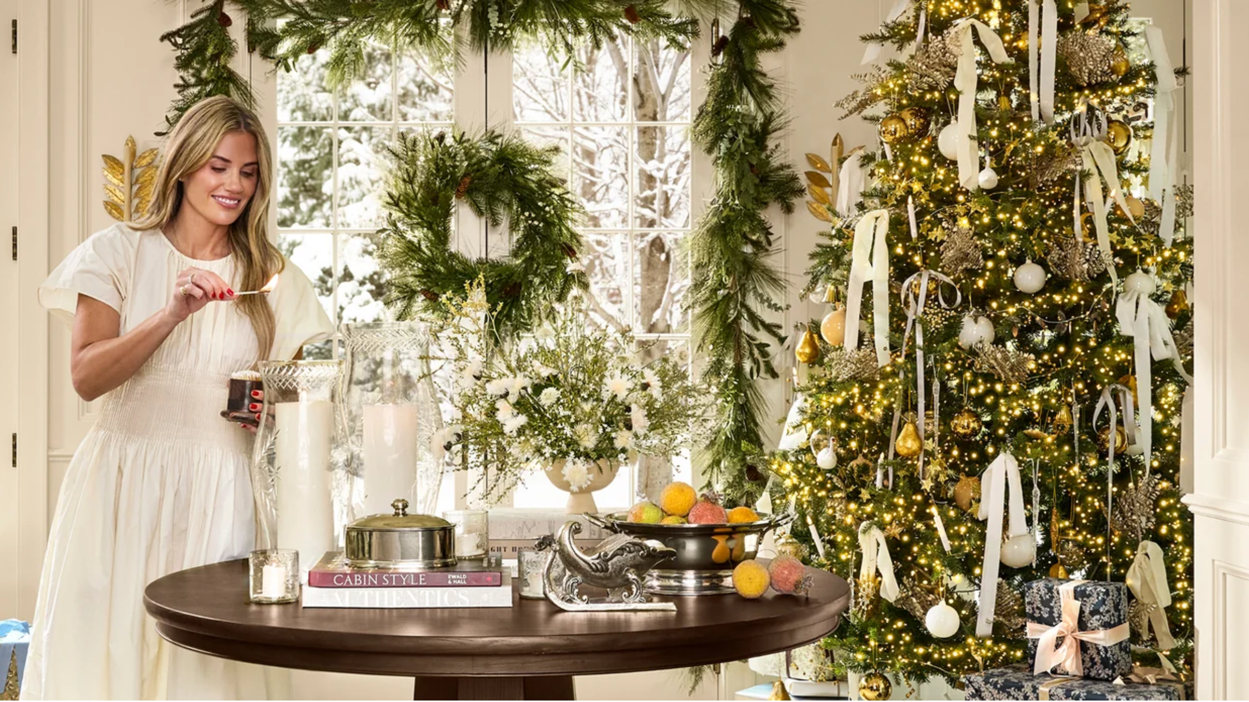 Shea McGee zündet eine große Stumpenkerze in einem festlichen Weihnachtswohnzimmer neben einem kunstvoll geschmückten Baum, einem Kranz, einer Girlande und metallischen Tischakzenten an.