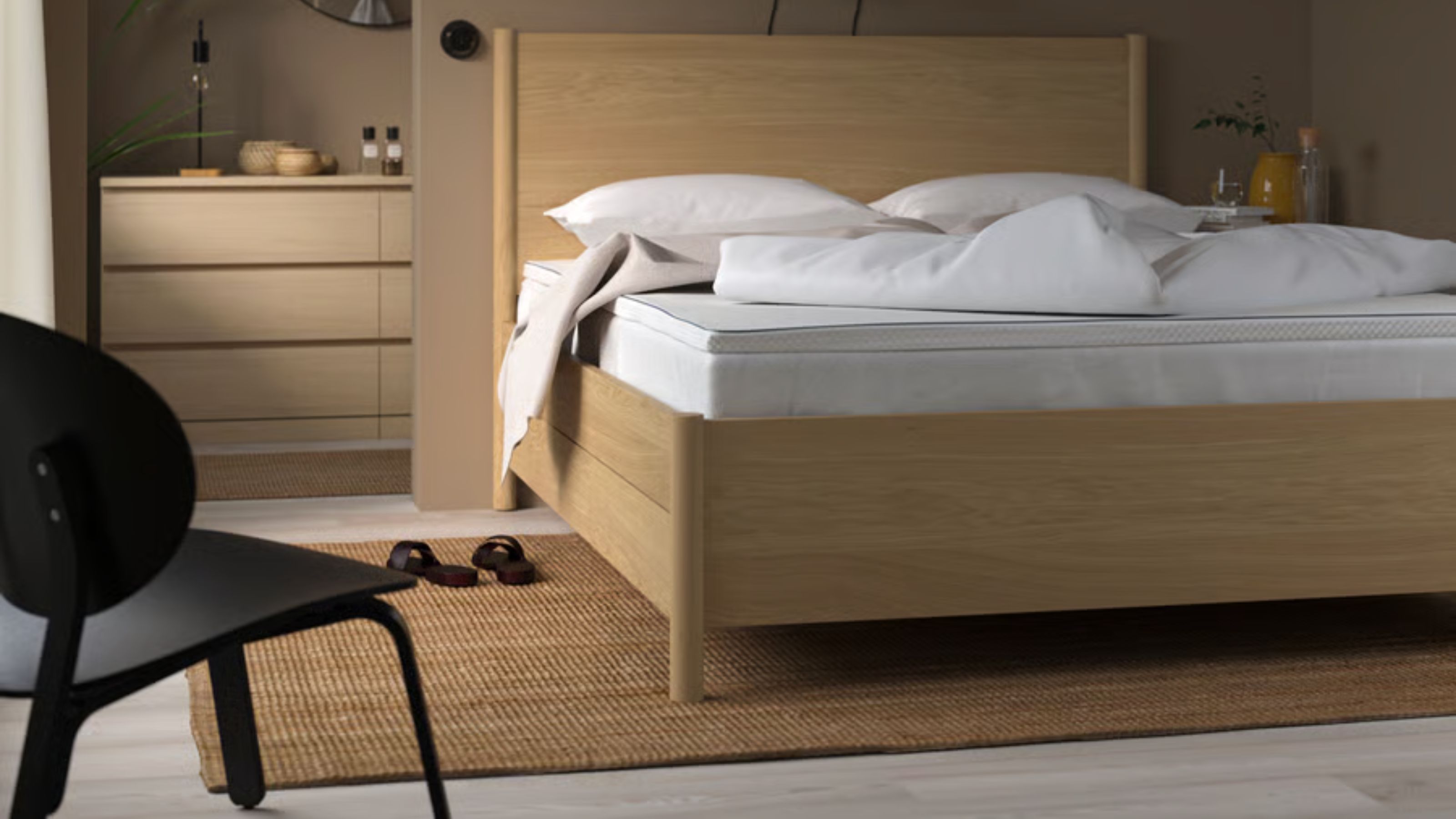 IKEA NÄSTABERG Matratzenauflage auf einem Doppelbett aus Kiefernholz in einem neutralen taupefarbenen Schlafzimmer mit schwarzem Stuhl, Juteteppich und Badezimmerschubladen und kreisförmigem Spiegel dahinter.