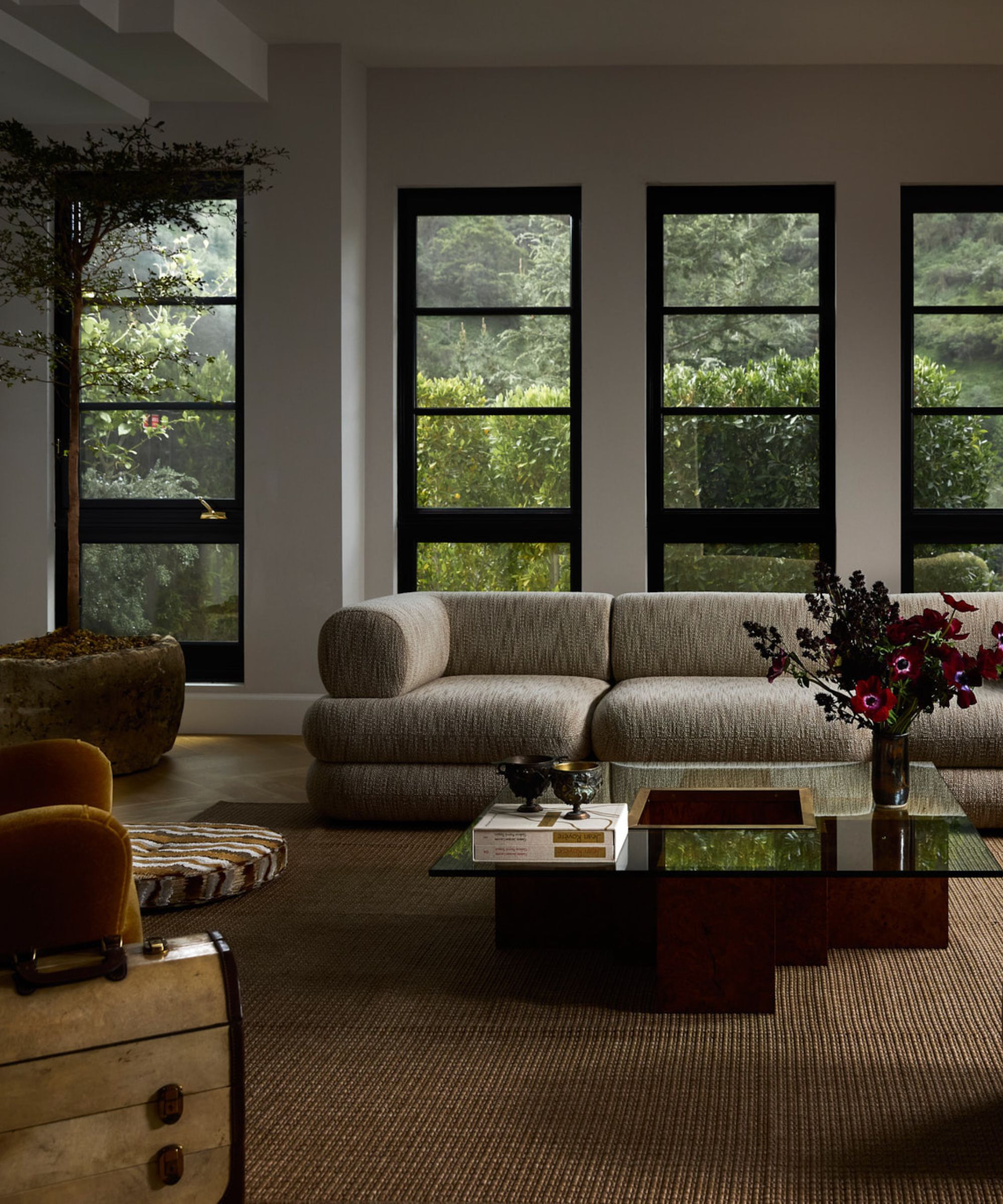 Großes, modernes Wohnzimmer, großes cremefarbenes Sofa mit gelben Sesseln und Couchtisch aus Glas und Holz, beiger Teppich und kleiner Ottomane, große Fenster mit viel Grün dahinter