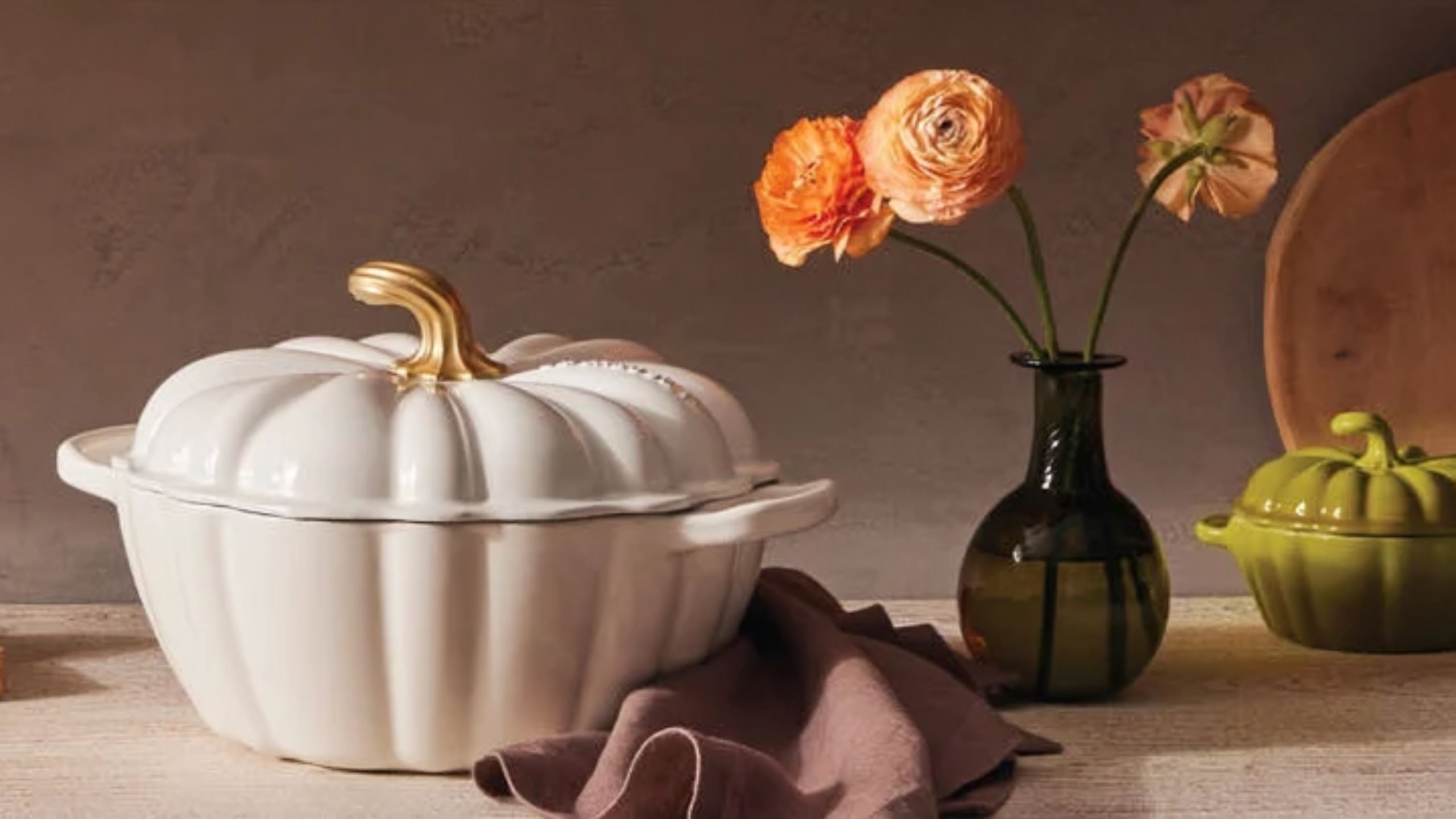 Eine weiße Le Creuset Pumpkin Cocotte auf einer Theke neben einer schwarzen Glasvase mit orangefarbenen Blumen. 