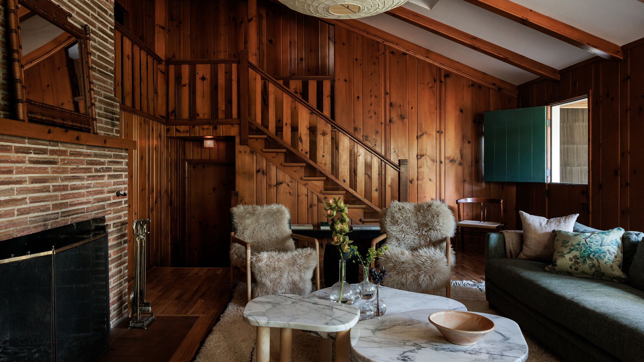 ein mit Holz verkleideter Hüttenwohnbereich mit gemauertem Kamin, Schaffellstühlen und drei Marmor-Couchtischen unter einem Globusanhänger