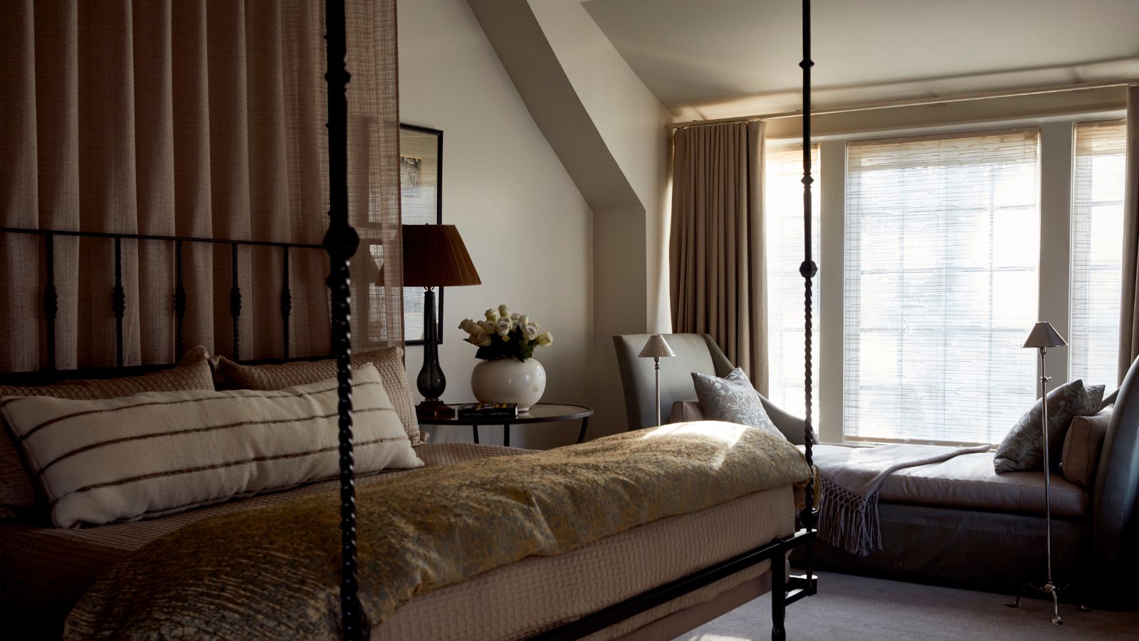 Ein gemütliches, neutrales Schlafzimmer mit warmweißen Wänden, einem großen Fenster mit Sitzbank und beigen Vorhängen, Teppichboden, einem Himmelbett mit Himmelbett und mehrlagiger neutraler Bettwäsche.