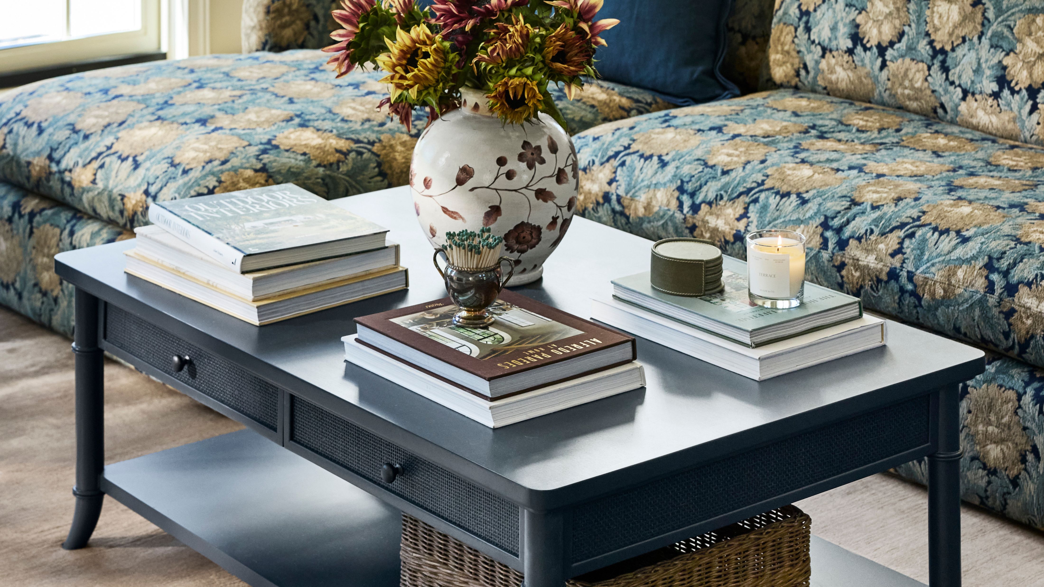 Zweistöckiger, marineblauer Couchtisch mit Stauraum, dekoriert mit Stapeln von Couchtischbüchern, einer Vase mit Blumenmuster, einem Streichholzanzünder und brennenden Kerzen.