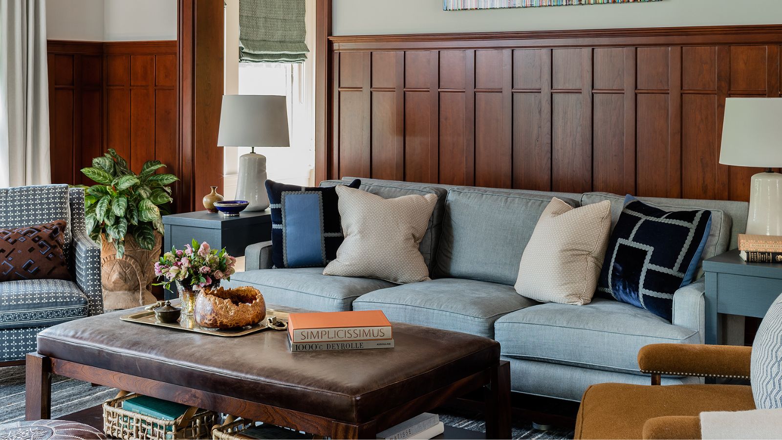 Wohnzimmer mit dunkelholzgetäfelten Wänden, einem hellblauen Sofa in der Mitte mit Kissen darauf, einem dunkelbraunen Ledercouchtisch, zwei Lederhockern darunter und einem senffarbenen Samtsessel.
