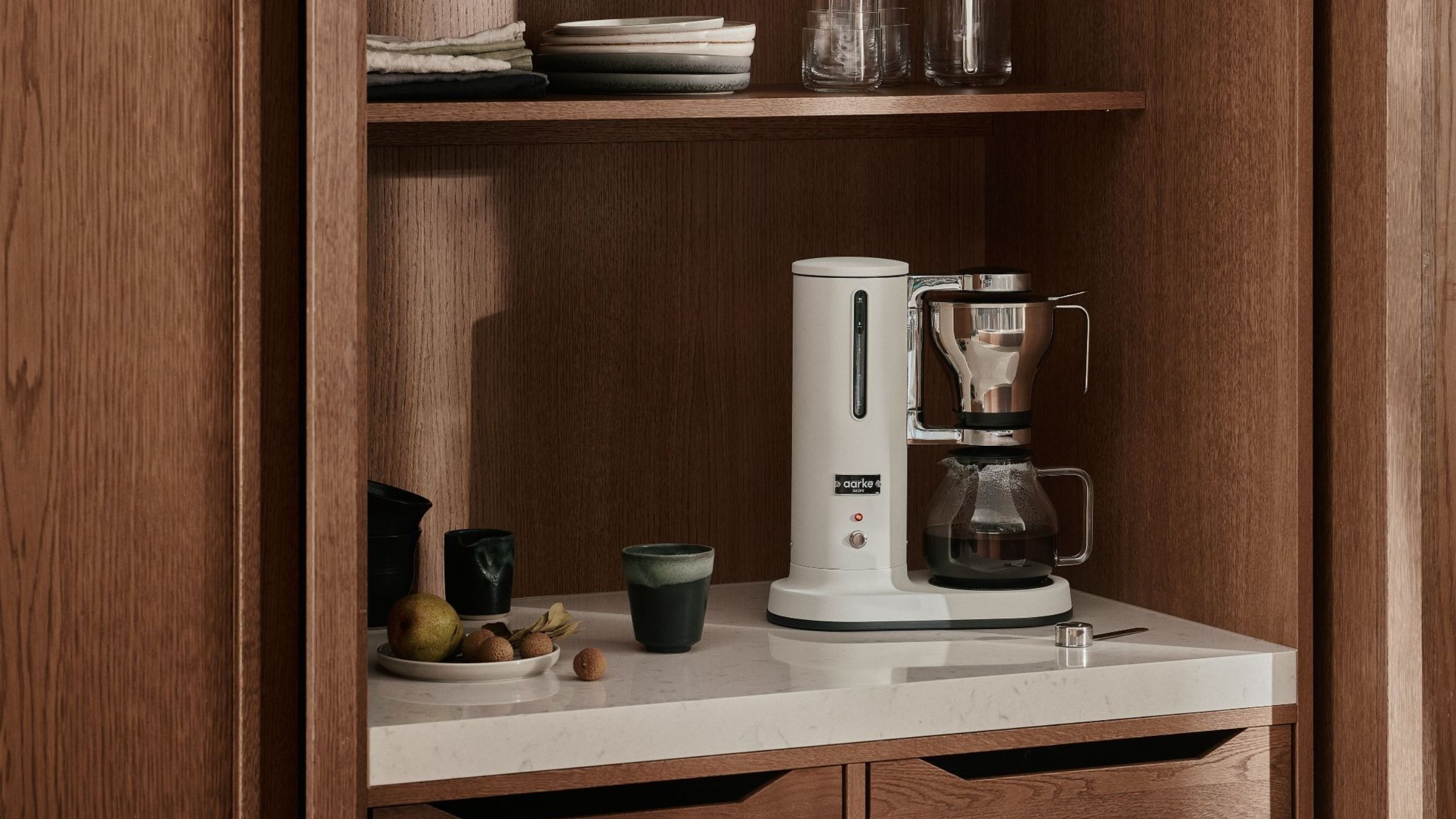 Eine weiße Filterkaffeemaschine von Aakre, versteckt in einer Gerätegarage aus dunklem Holz mit weißen Arbeitsflächen. 