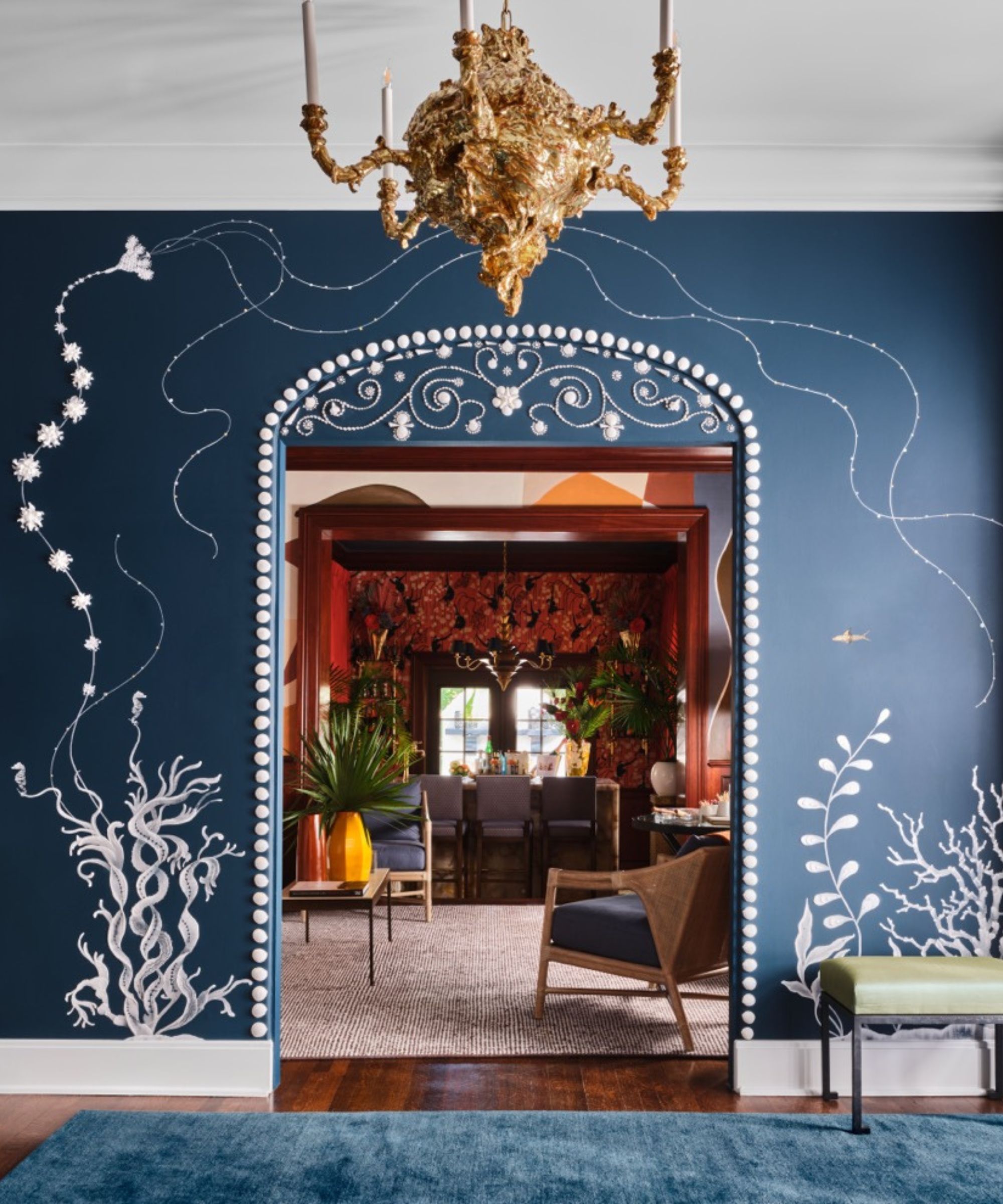 Blauer Eingangsbereich mit blau gestrichenen Wänden und weißen Blumengemälden, Blick durch und Torbogen in ein orangefarbenes Wohnzimmer mit Möbeln und einem großen neutralen Teppich