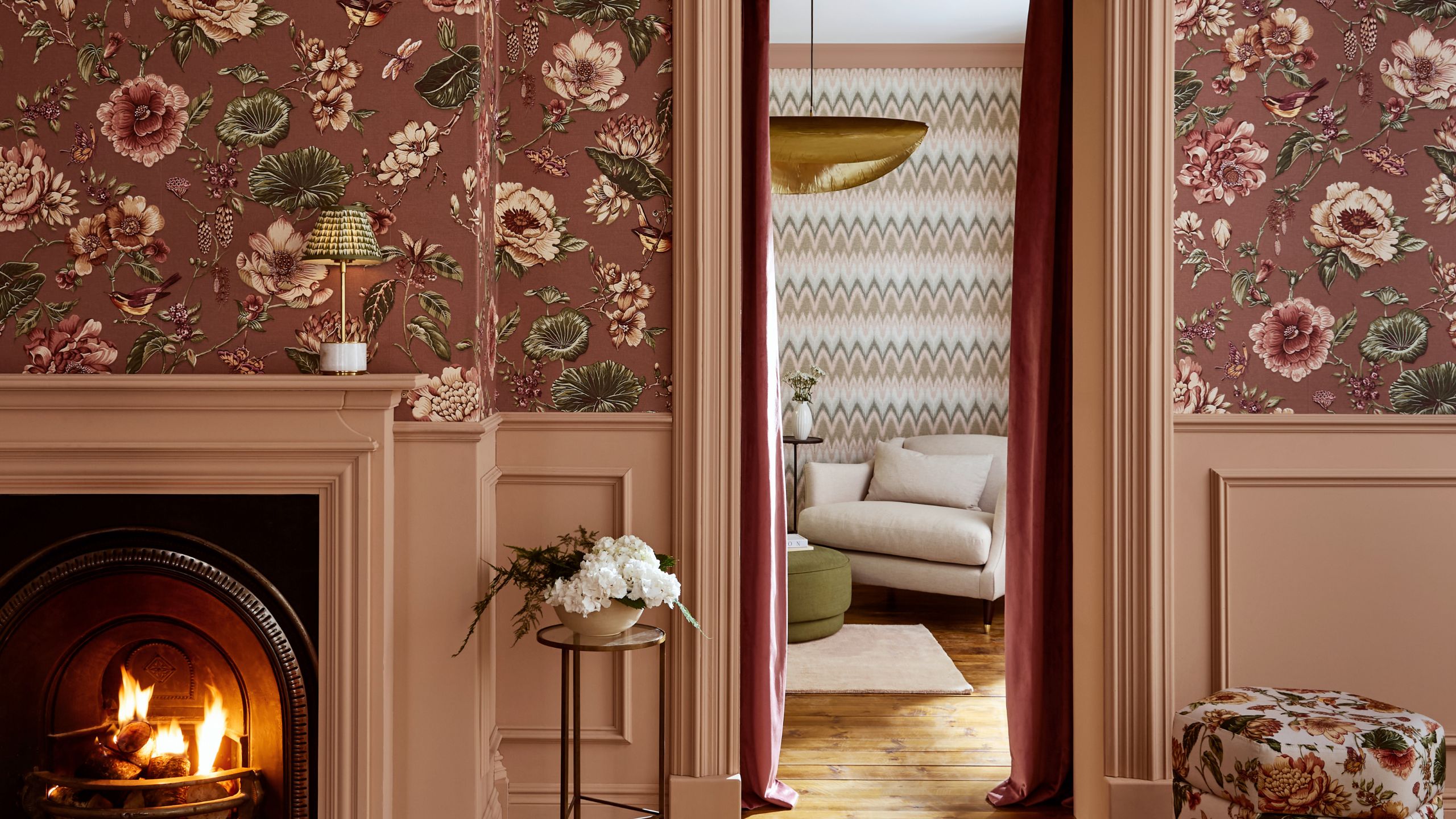 Wohnzimmer mit rosafarbener Blumentapete, altrosafarbener Täfelung, Kamin, geblümter Ottomane, Blick in ein weiteres Wohnzimmer mit Chevron-Tapete und cremefarbenem Sessel.