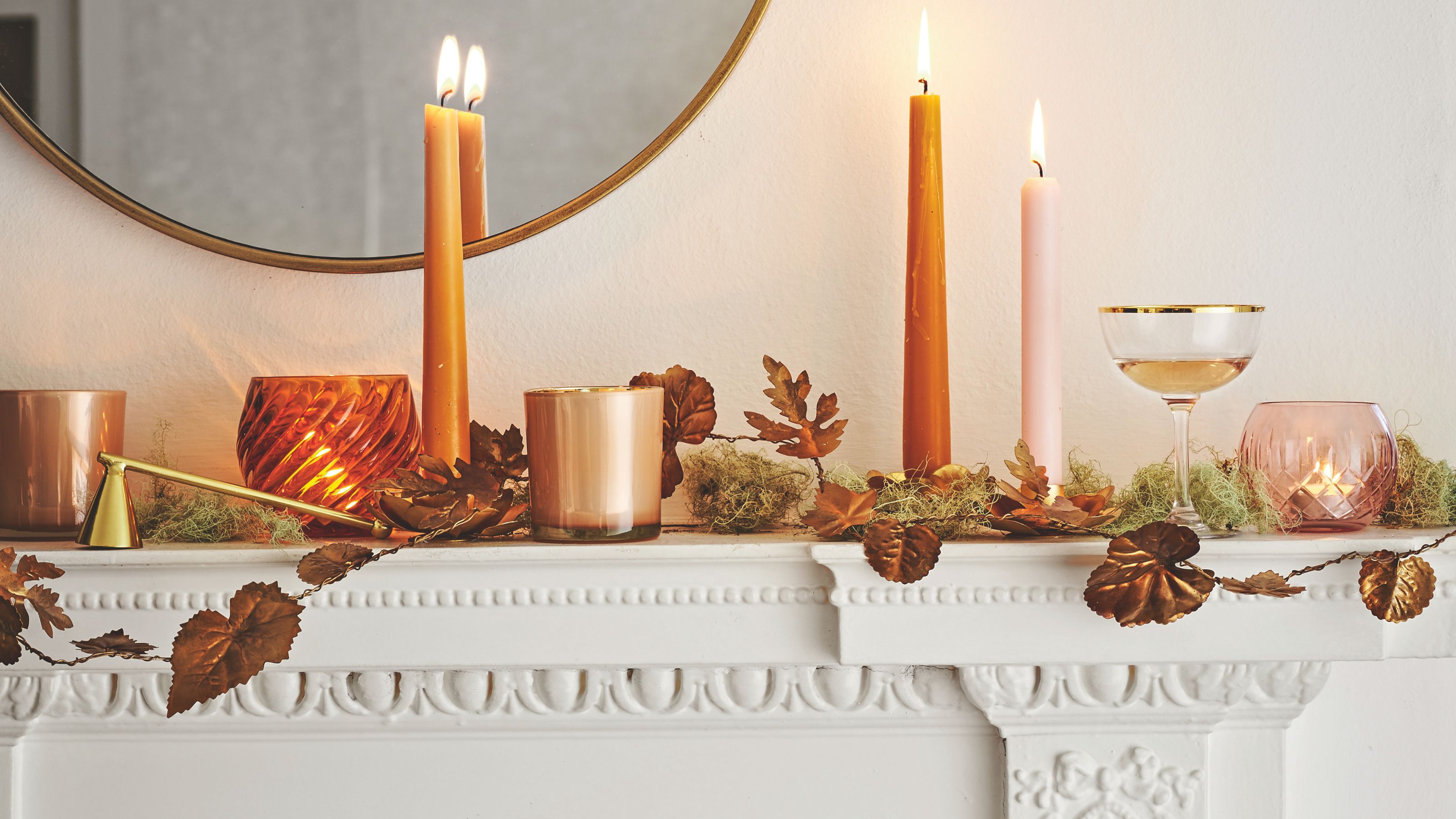 Weißer herbstlicher Kaminsims mit brennenden orangefarbenen Stabkerzen, Blättergirlande und rundem Spiegel