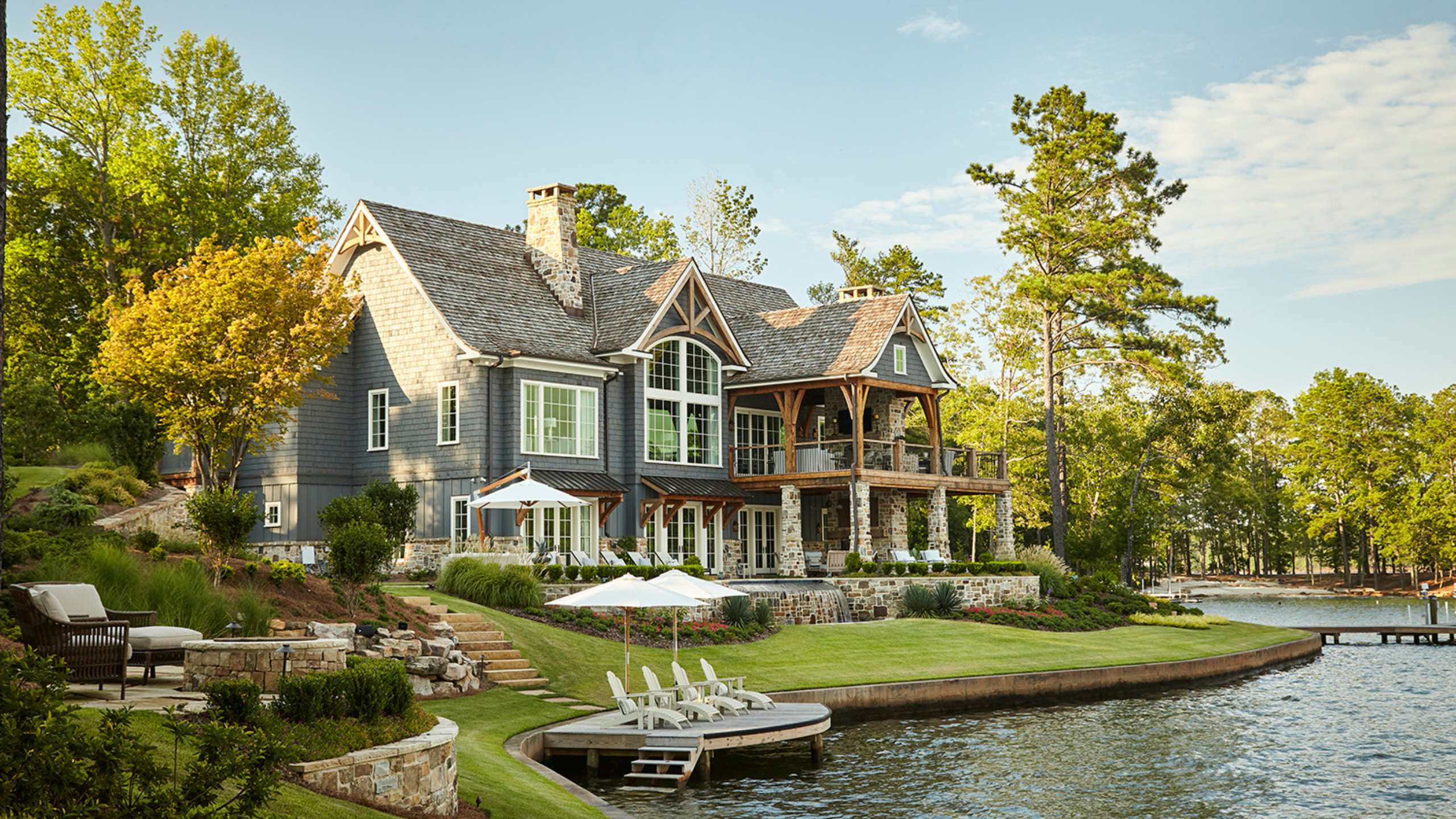 Grand waterfront home on Lake Martin featuring rows of chaise lounges shaded by bright white umbrellas. A tall forest of trees surrounds the scene.