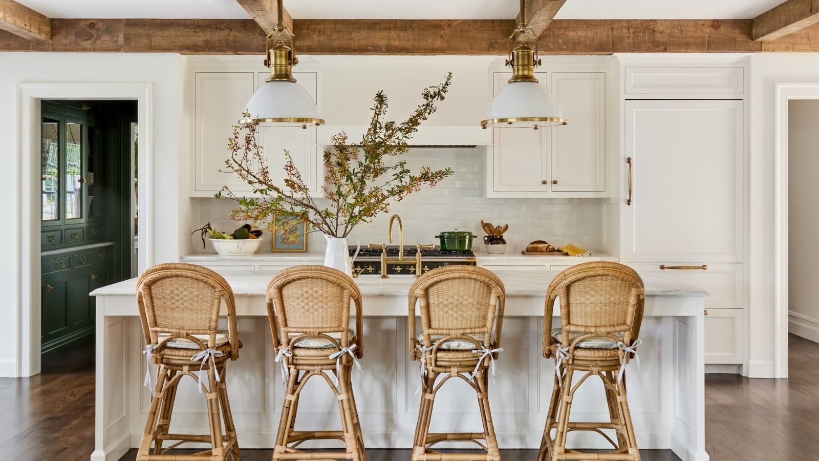 A white kitchen with wooden ceiling beams, rattan bar stools, and fall foliage displayed in a vase on the island