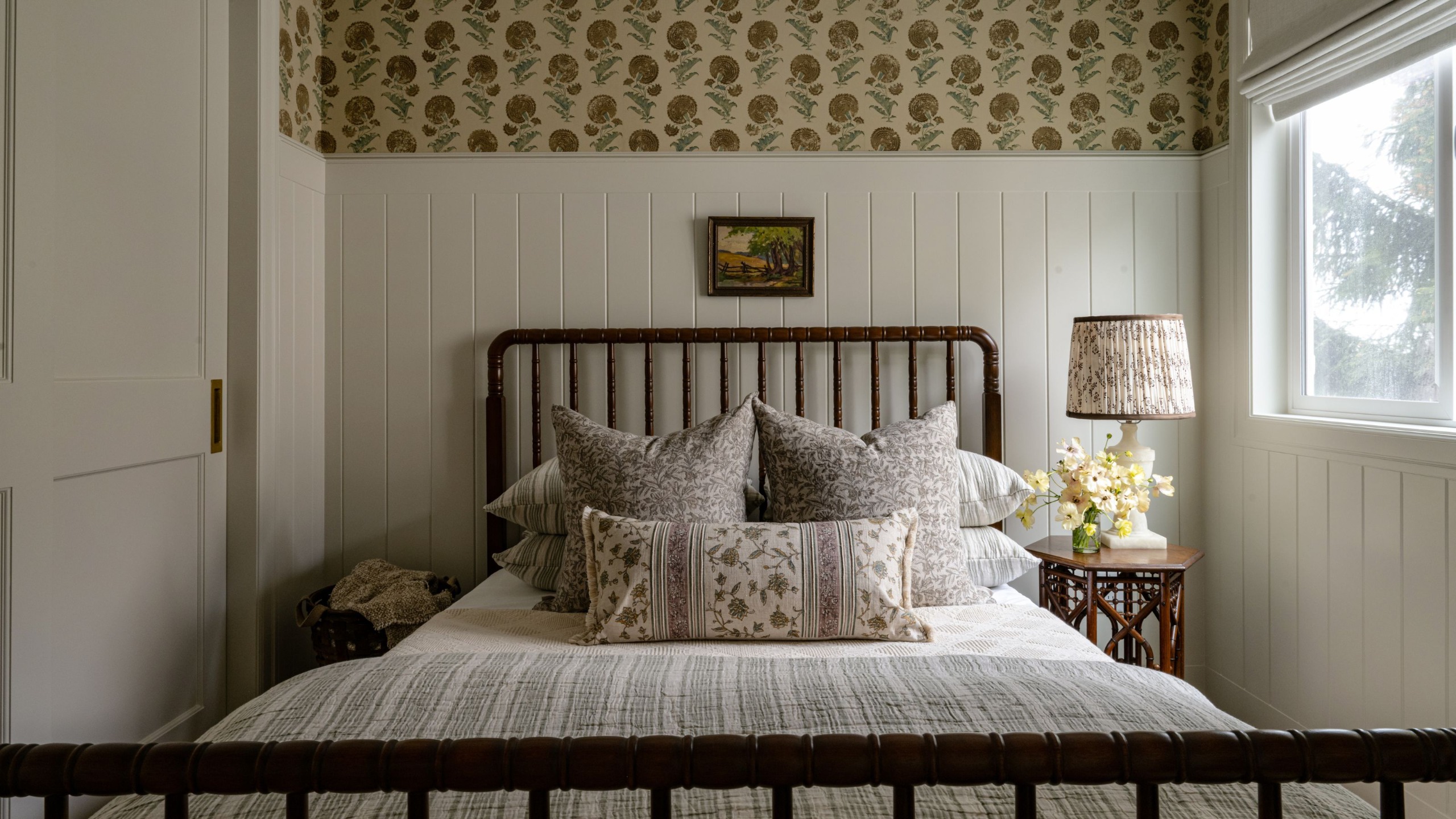 Ein gemütliches Gästezimmer mit warmweißer Täfelung, kleinen Blumentapeten an den oberen Wänden und Einbauschränken. Ein Spindelbettgestell aus dunklem Holz mit neutral gemusterter Bettwäsche und einer gemusterten Tischlampe. 