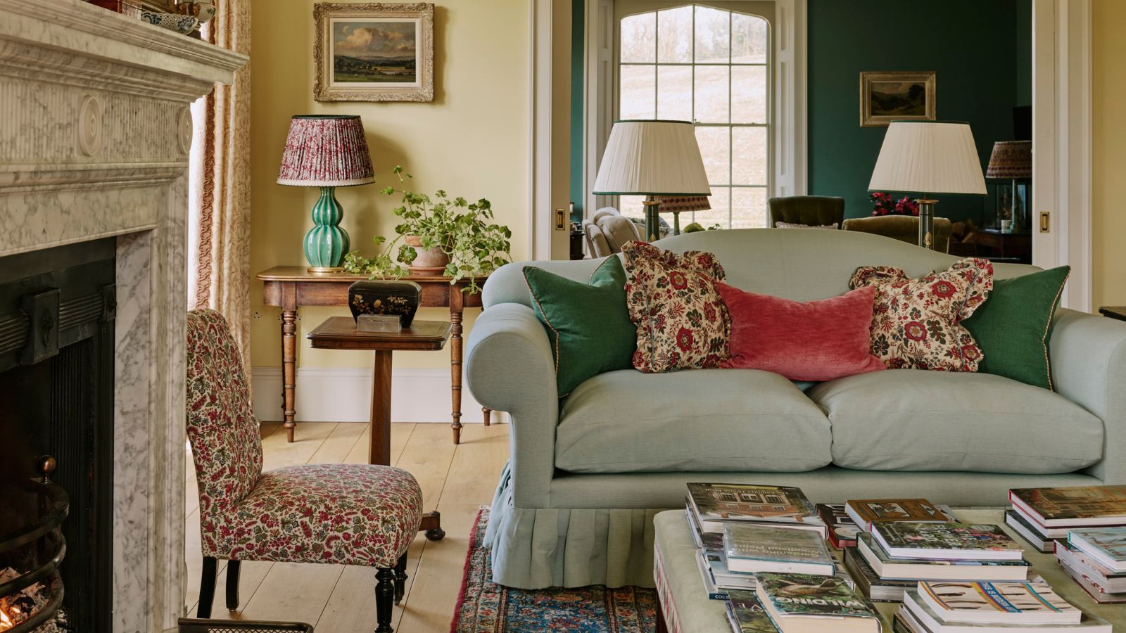 Elegantly styled living room featuring a light blue skirted sofa with colorful accent pillows, a marble fireplace, an upholstered coffee table stacked with books, and an doorway opening into a teal room.