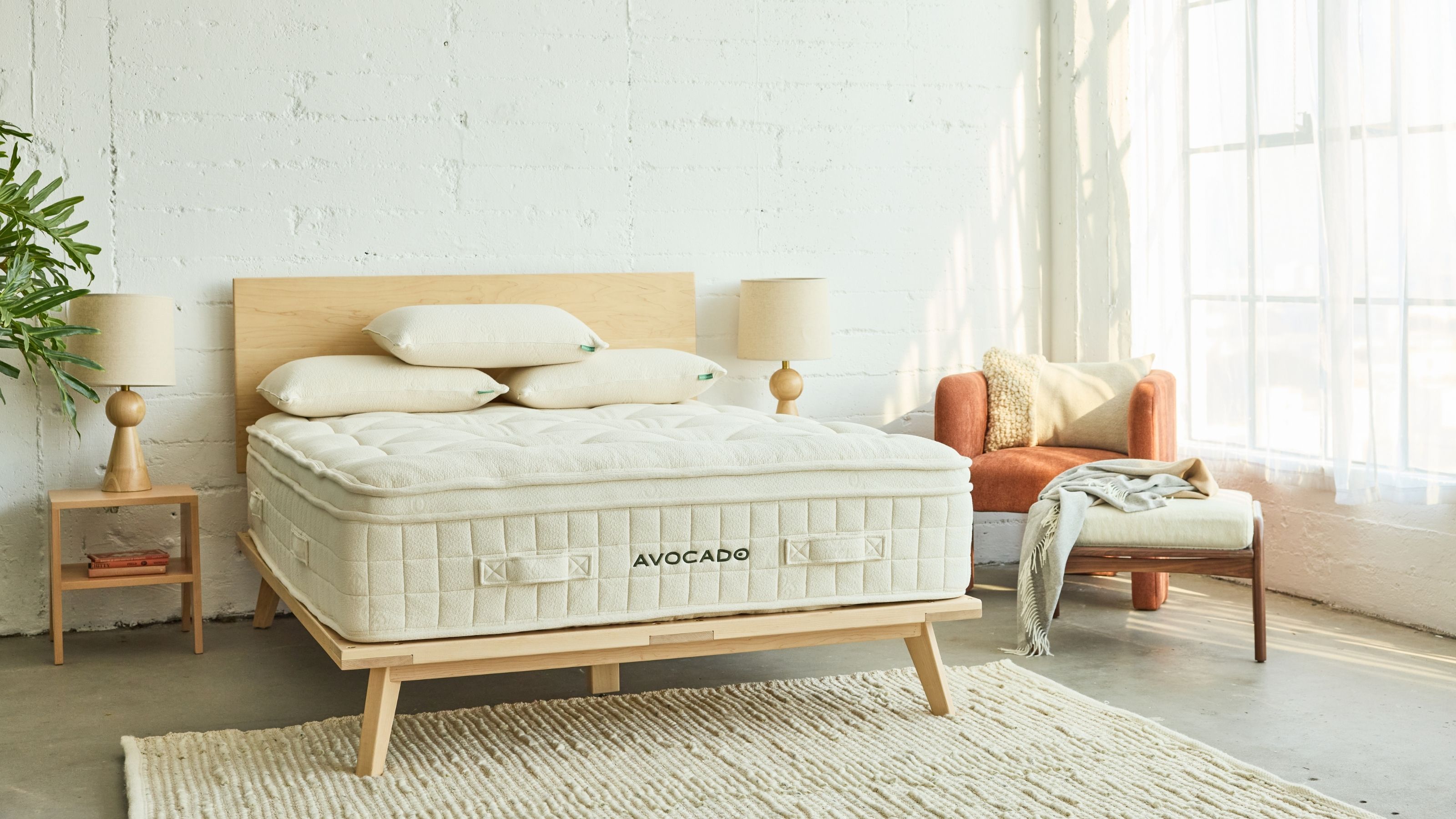 Avocado-Luxus-Bio-Matratze auf einem hellen Holzbettgestell in einem weißen Schlafzimmer mit Holzboden, freiliegenden, weiß gestrichenen Ziegelwänden und großen Fenstern auf der rechten Seite.