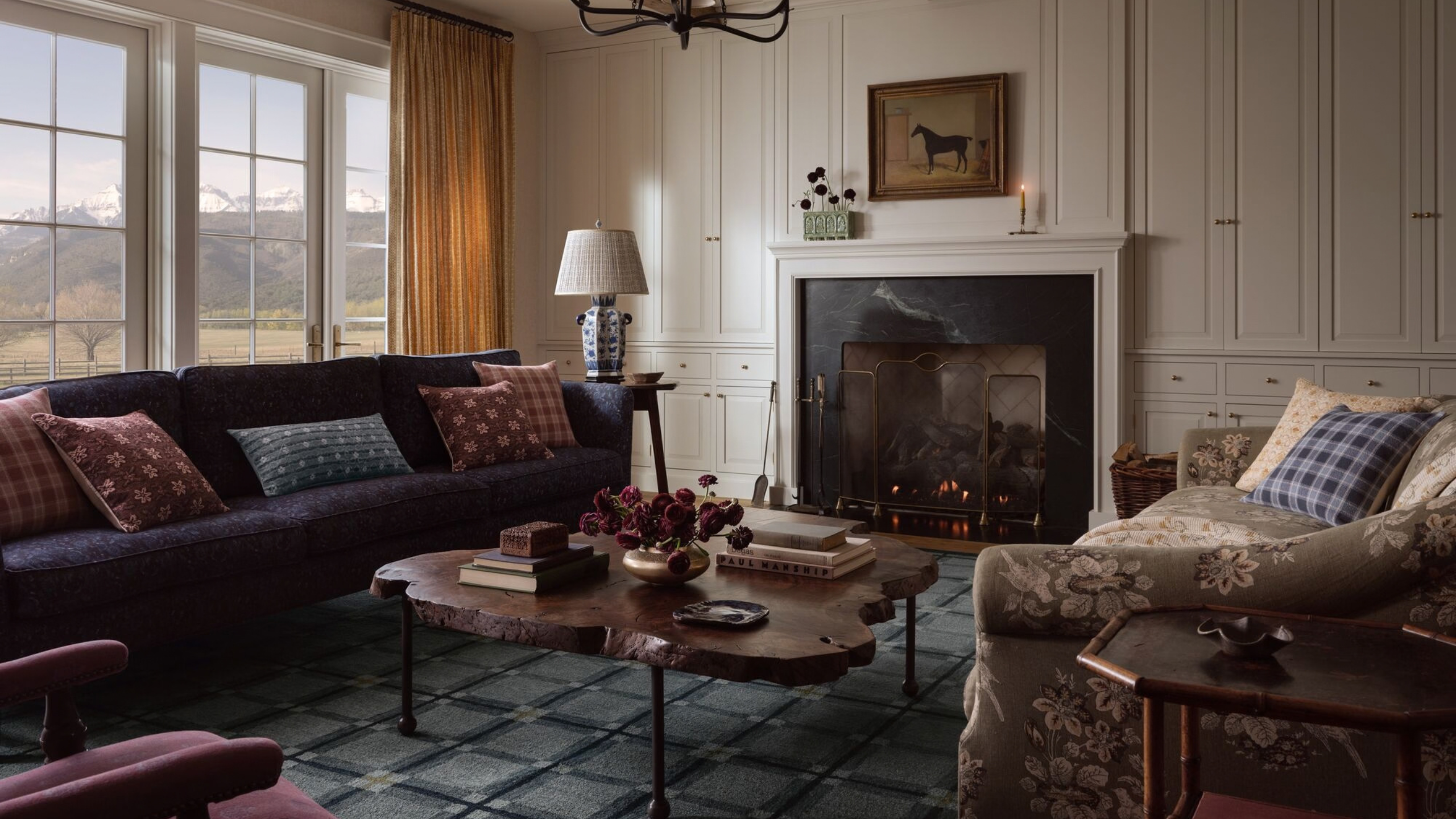 Stimmungsvolles Wohnzimmer mit einem blauen geometrischen Teppich, einem marineblauen Sofa, einem Zweiersofa mit Blumenmuster, einem Couchtisch aus Echtholz, beigen Vorhängen, Kissen im Vintage-Stil und Reitkunst über dem Kamin
