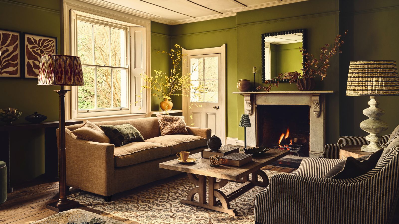 Ein tiefgrünes Wohnzimmer mit zwei Sofas und einem Holztisch in der Mitte. Ein großer Steinkamin mit brennendem Feuer im Inneren. 