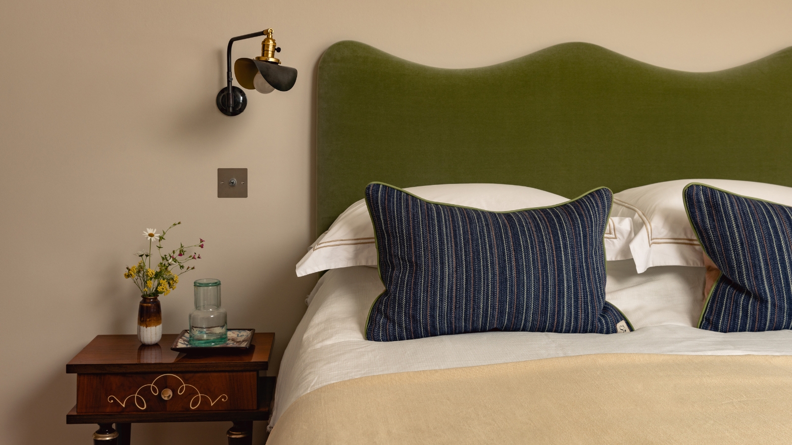 Schlafzimmer mit warmen, neutralen Wänden, einem Kopfteil aus grünem Samt, gestreiften marineblauen Kissen und einem Nachttisch aus dunklem Holz.