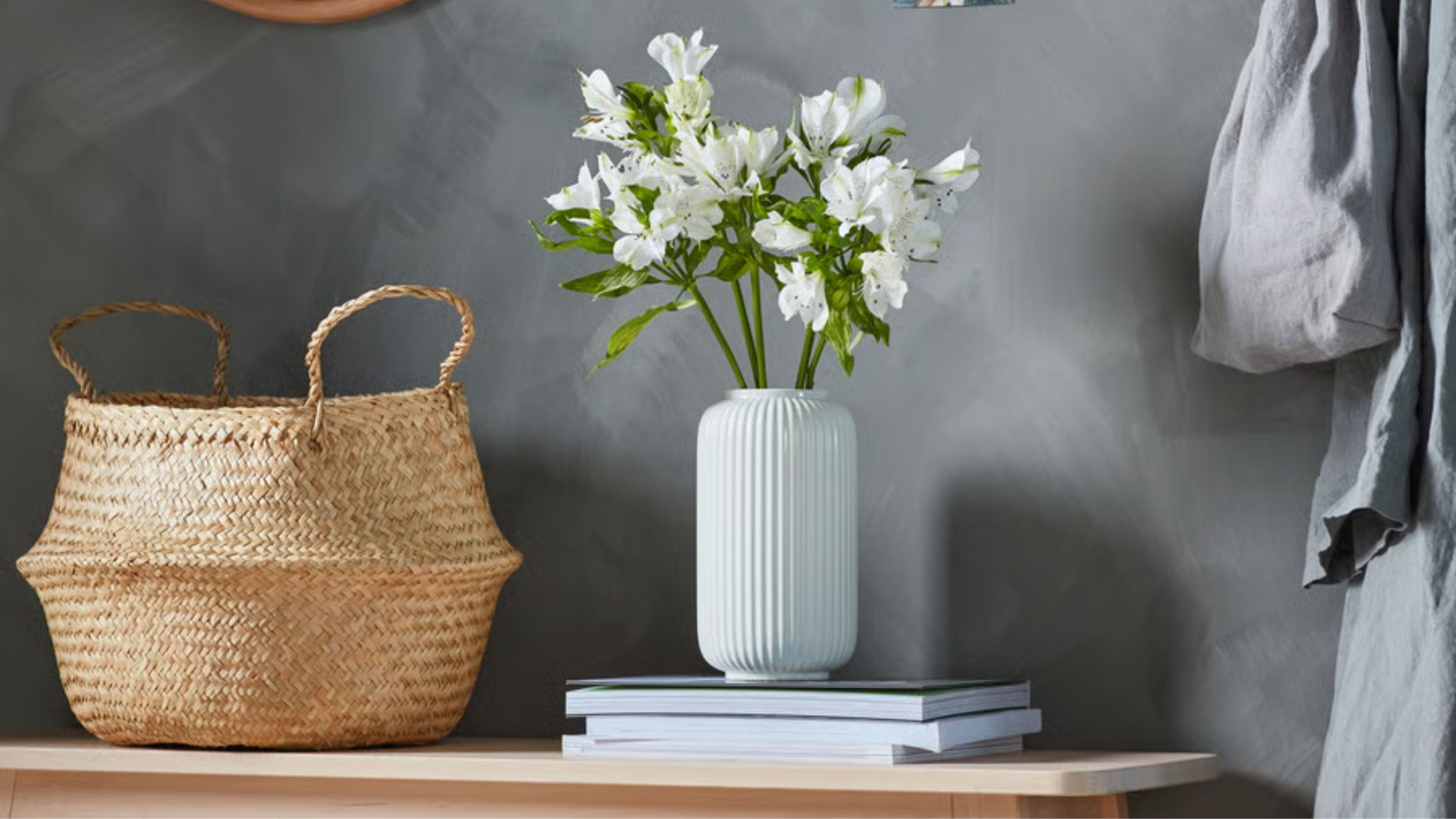 IKEA FLÅDIS Korb vor einer dunkelgrauen Wand neben einem Stapel Zeitschriften mit einer weißen geriffelten Vase voller weißer Blumen.
