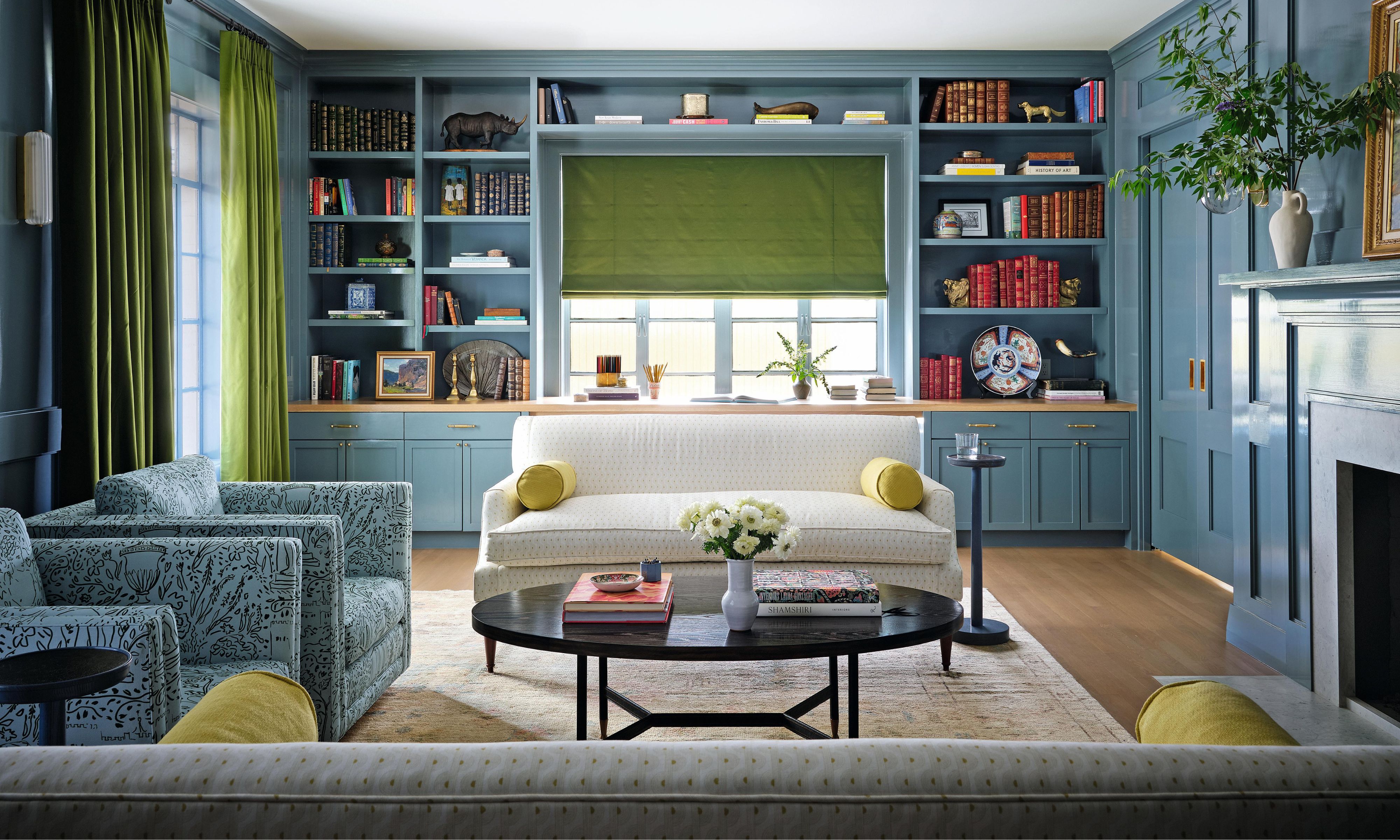 ein blau gestrichenes Wohnzimmer mit maßgefertigten Bücherregalen und Stauraum rund um das Fenster mit einer weißen Couch, zwei blau gemusterten Sesseln und einem schwarzen runden Couchtisch
