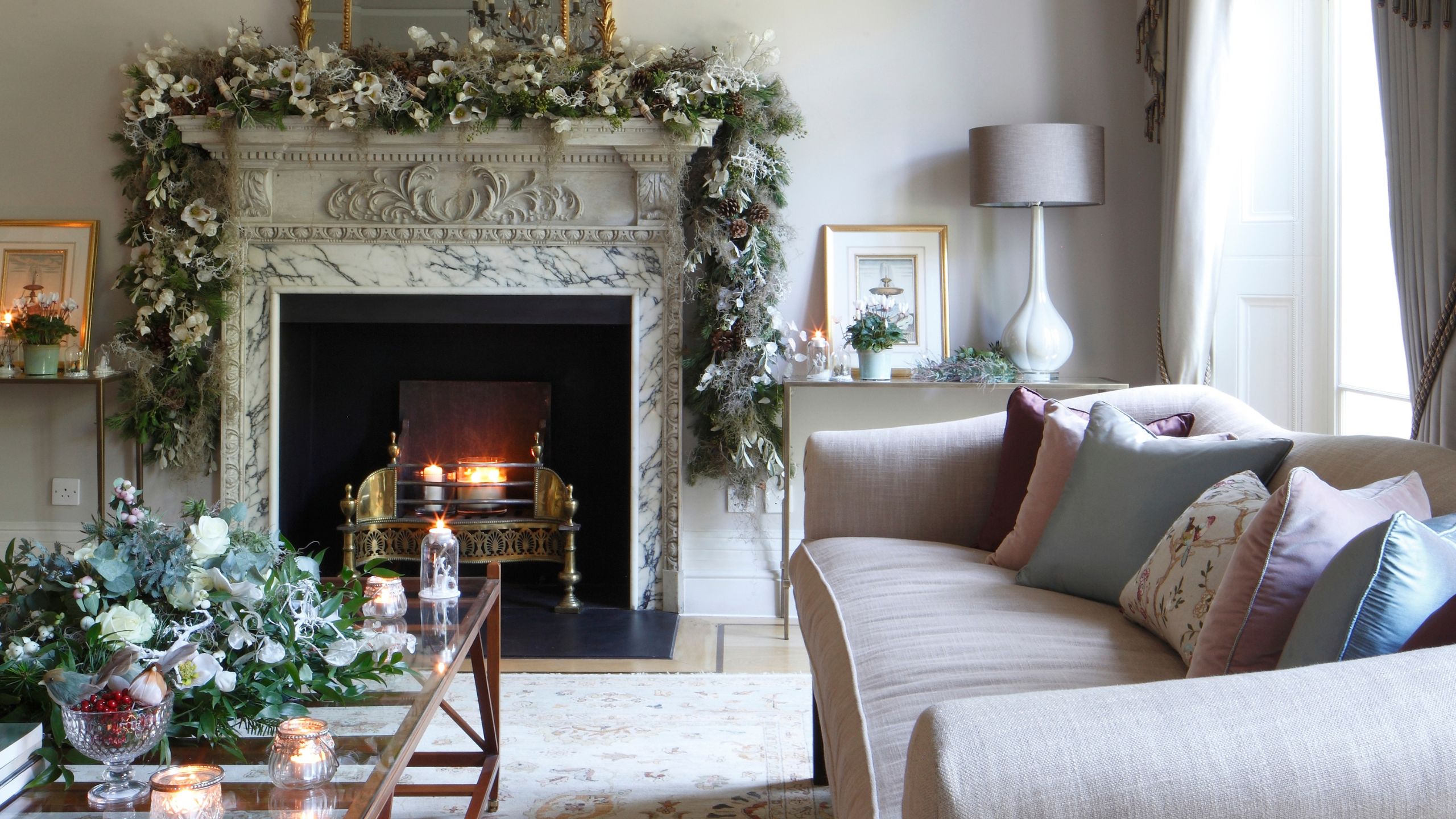 Neutrales Wohnzimmer mit Marmorkamin und brennendem Feuer, rosa Couch, Weihnachtsgirlanden, Couchtisch aus Glas und brennenden Kerzen.