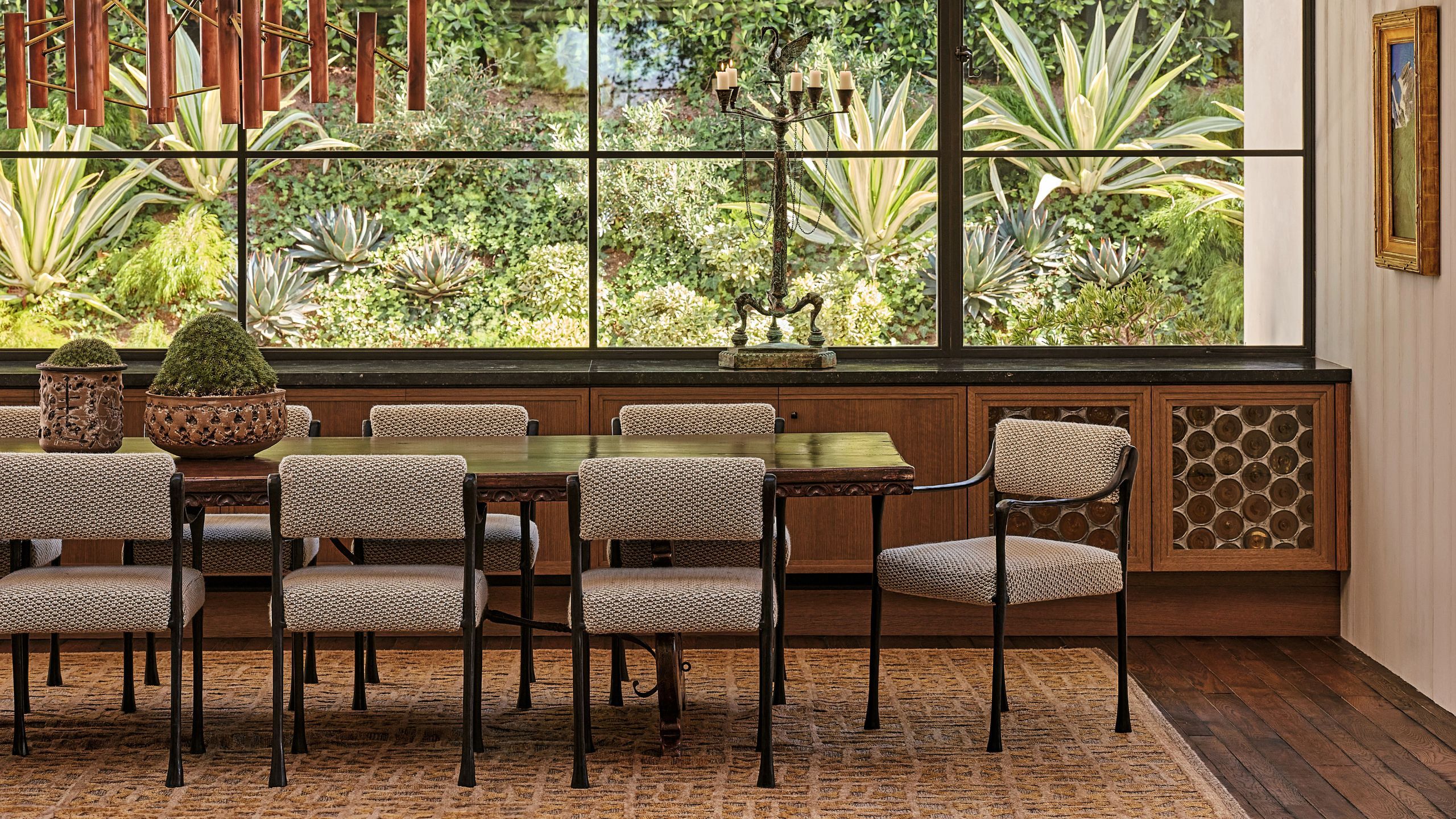 Mid-century dining table and woven chairs facing a large black-framed picture window looking out onto lush agave plants and garden greenery