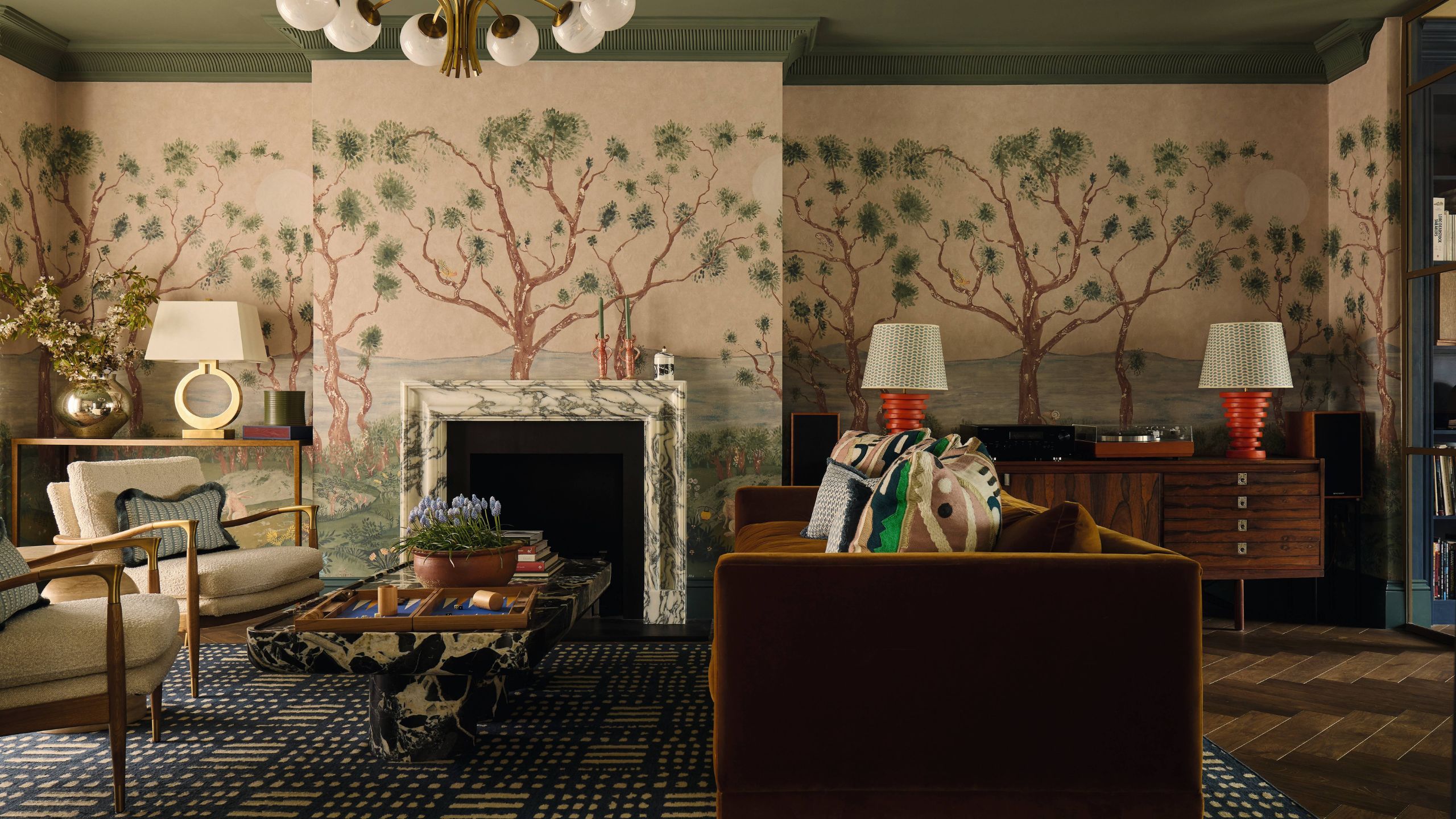 Ein großes Londoner Wohnzimmer mit rosa Wandgemälden und Baummotiven, die die Wände und den Kaminsims bedecken, einer grün gestrichenen Decke, einem Marmorkamin und vielseitigen Möbeln rund um ein toffeefarbenes Samtsofa