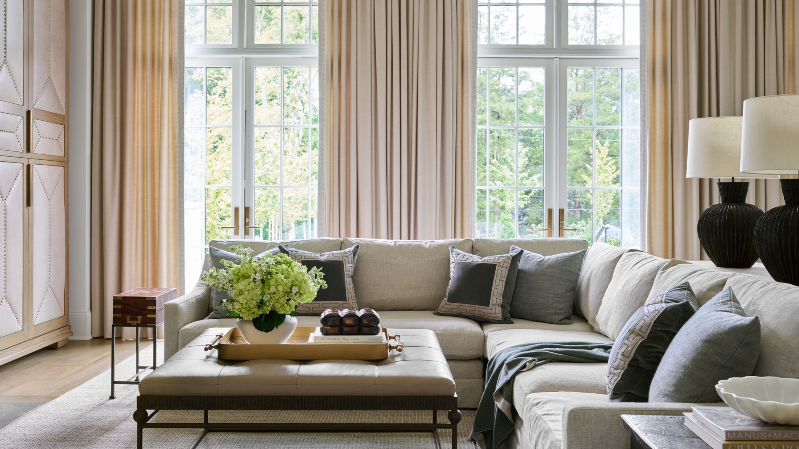 Modernes Wohnzimmer mit hohen Decken und Glastüren, die in den Garten führen, langen cremefarbenen Vorhängen, einem großen cremefarbenen Sofagarnitur, grauen Kissen, einem Couchtisch aus Leder und Metall mit Dekor darauf.