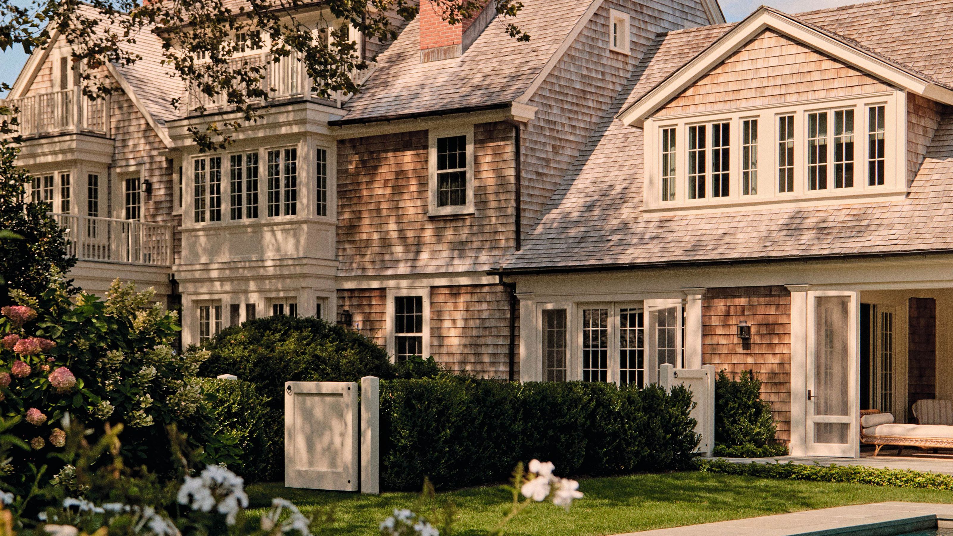 Das Äußere eines riesigen Backsteinhauses in Hamptons mit sanften beigen Außenverkleidungen und einem üppig grünen Poolgarten