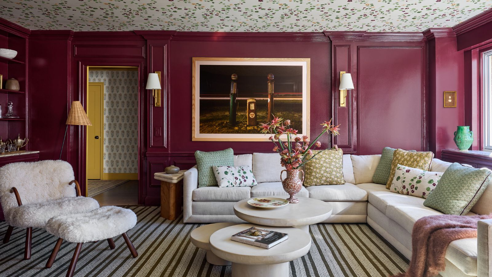 Ein Wohnzimmer mit burgunderrot getäfelten Wänden in Glanzoptik und einer Decke mit Blumentapete. Ein gestreifter Teppich, ein großes cremefarbenes Sofa mit grünen, gelben und geblümten Kissen, ein moderner dreiteiliger Couchtisch, Kunstwerke an der Wand mit zwei Wandleuchten und ein Akzentstuhl aus Lammfell. 