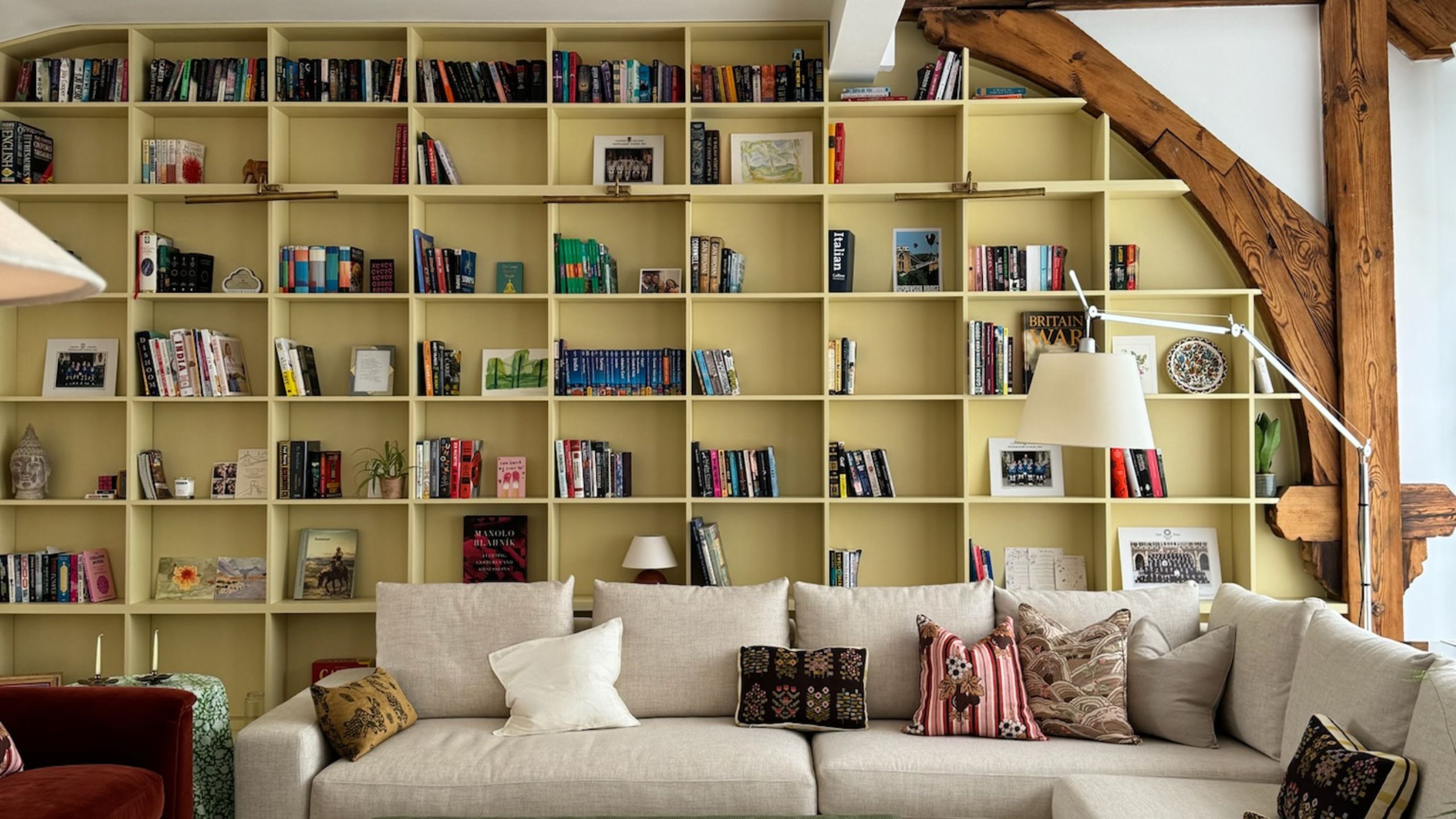Wohnzimmer mit Balkendecke und -wänden sowie einem großen eingebauten, gelb gestrichenen Bücherregal, gefüllt mit Büchern in jeder Nische. In der Mitte steht ein großes cremefarbenes Sofagarnitur mit gemusterten Kissen.