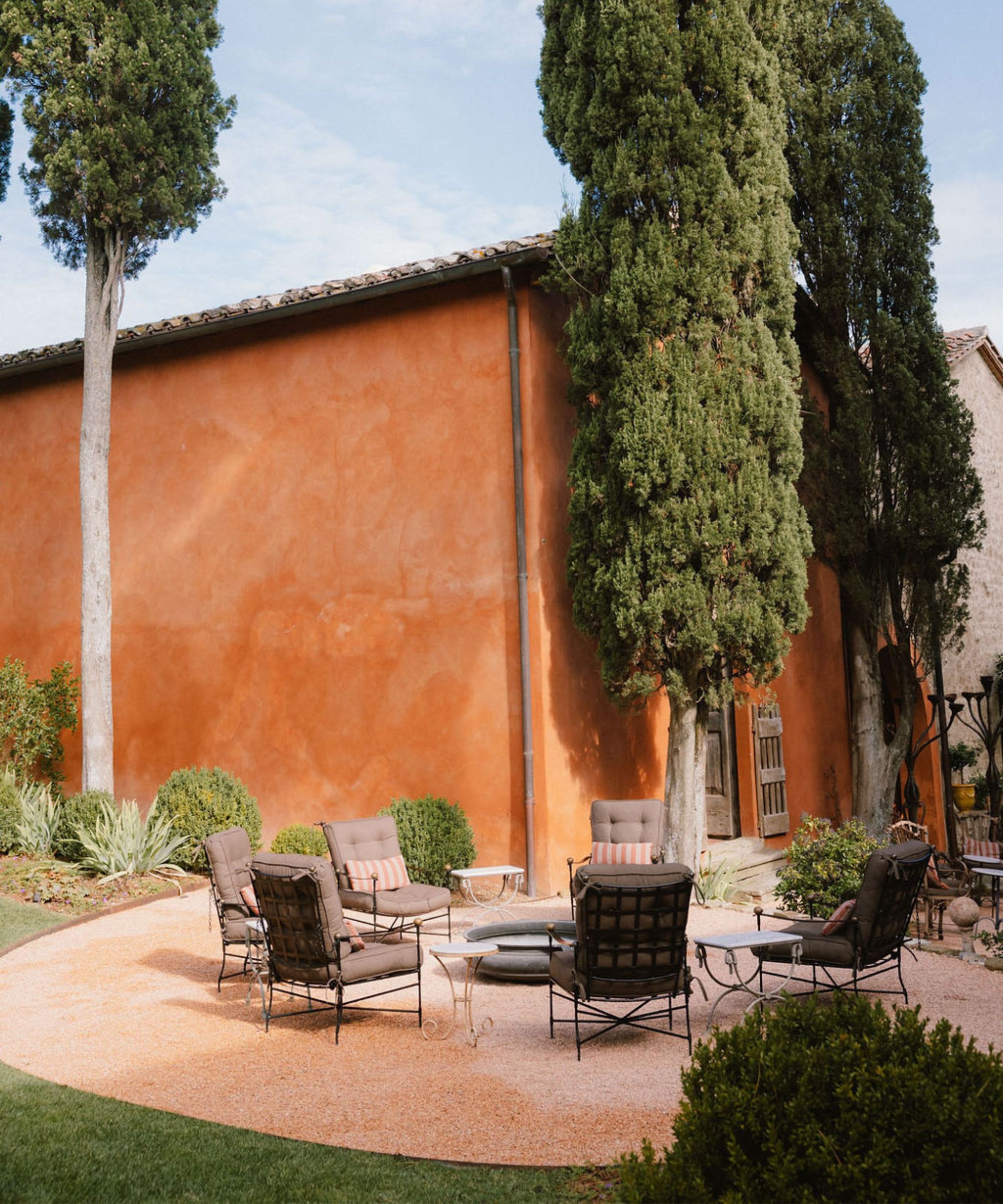 Italienische Terrasse mit Terrakotta-bemaltem Gebäude, umgeben von Bäumen und Pflanzen. Sitzbereich auf der Steinterrasse rund um die Feuerstelle